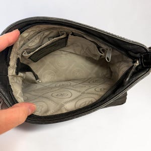May include: The interior of a dark gray handbag featuring a light beige fabric lining with a repeating circular logo pattern. The bag includes a zippered main compartment, an internal zippered pocket, and an open slip pocket for organization.