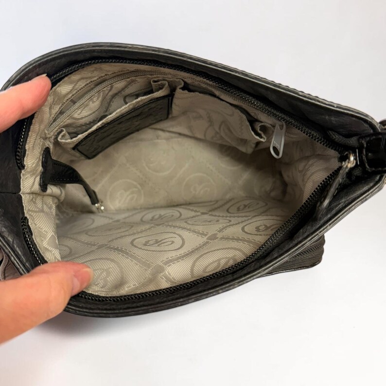 May include: The interior of a dark gray handbag featuring a light beige fabric lining with a repeating circular logo pattern. The bag includes a zippered main compartment, an internal zippered pocket, and an open slip pocket for organization.