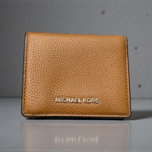May include: A small, rectangular, tan leather wallet with a textured surface. The wallet has the words "MICHAEL KORS" in silver lettering on the front. The wallet is closed and appears to be new.