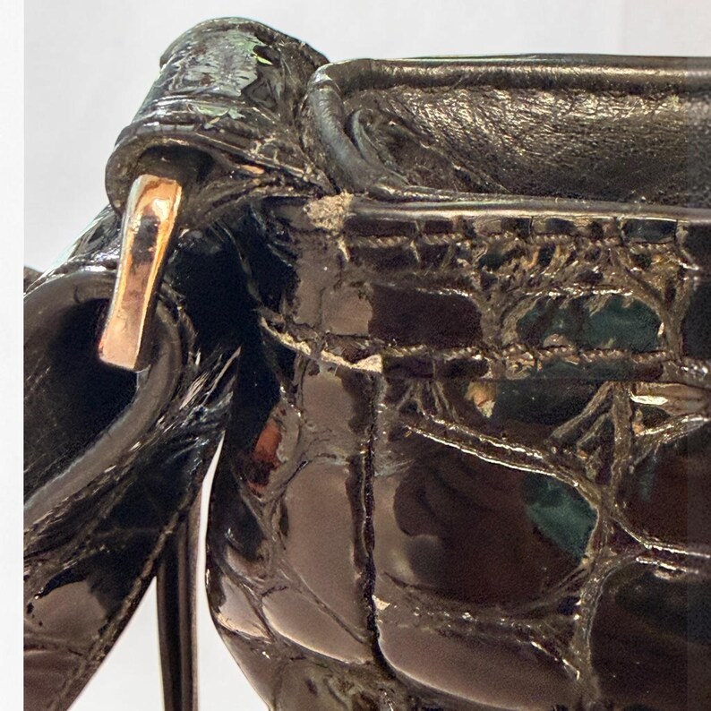 May include: A black patent leather handbag featuring a glossy crocodile-embossed texture. The bag includes a metallic silver-tone hardware attachment for the strap, visible stitching along the top edge, and signs of wear on the upper trim.