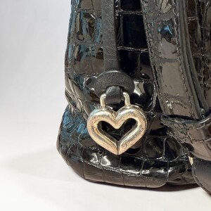 May include: A black handbag with a glossy, embossed crocodile-skin texture. A silver-toned metal heart-shaped charm hangs from a black leather strap, featuring the engraved brand name Brighton on the lower right side of the heart.