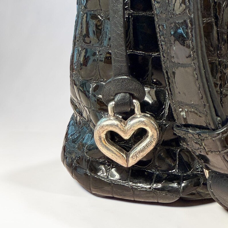 May include: A black handbag with a glossy, embossed crocodile-skin texture. A silver-toned metal heart-shaped charm hangs from a black leather strap, featuring the engraved brand name Brighton on the lower right side of the heart.