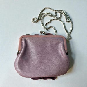 Puede incluir: Un monedero de cuero de color lavanda con una correa de cadena plateada. El bolso tiene un cierre de beso y un detalle floral decorativo en la base. La cadena es de dise&ntilde;o delicado y tejido.