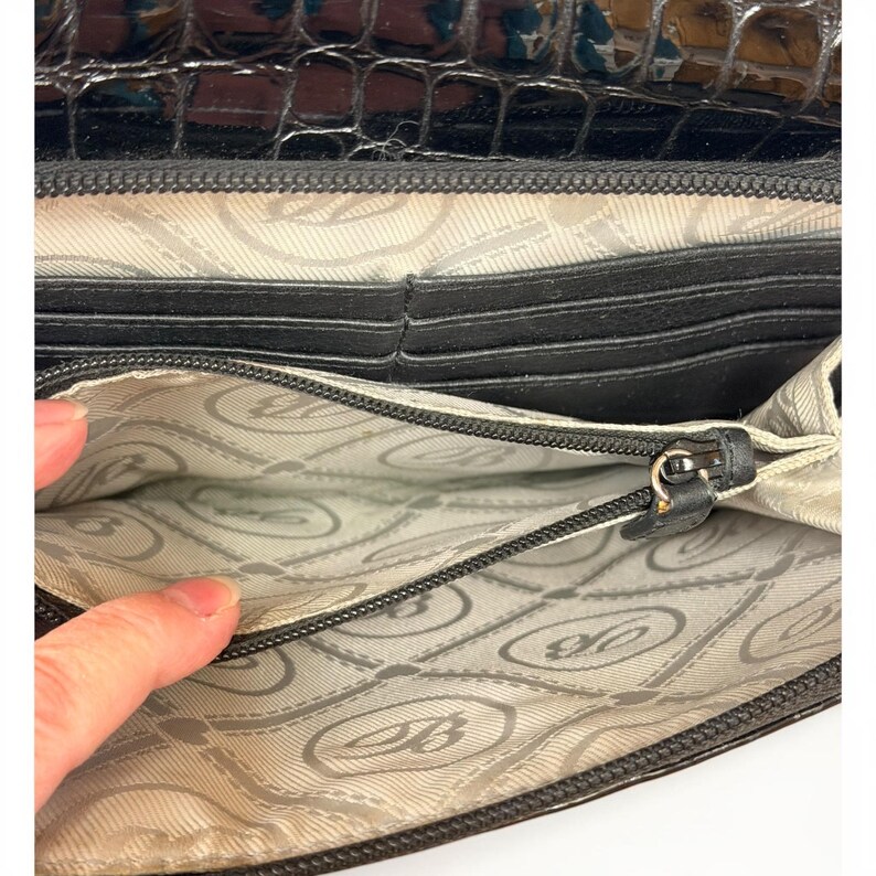 May include: A black wallet with a glossy crocodile-embossed exterior and a light gray interior lining featuring a repeating circular logo pattern. The interior includes multiple card slots and a central zippered compartment with a black zipper pull.