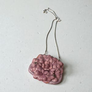 Puede incluir: Un collar con colgante en forma de flor rosa. El colgante est&aacute; hecho de p&eacute;talos en capas y tiene una cadena plateada. El collar est&aacute; sobre un fondo blanco.