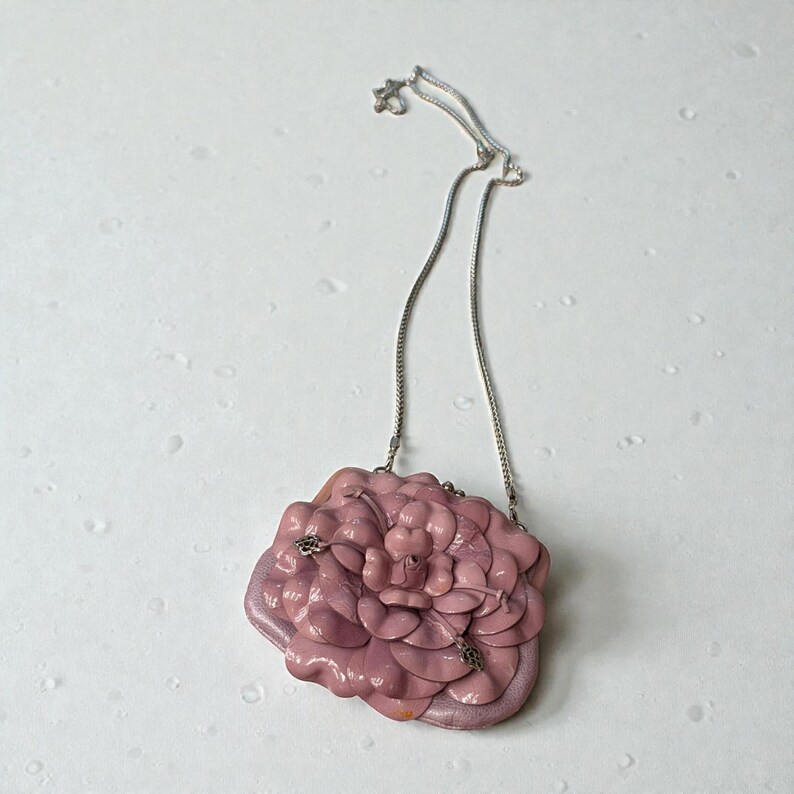 Puede incluir: Un collar con colgante en forma de flor rosa. El colgante est&aacute; hecho de p&eacute;talos en capas y tiene una cadena plateada. El collar est&aacute; sobre un fondo blanco.