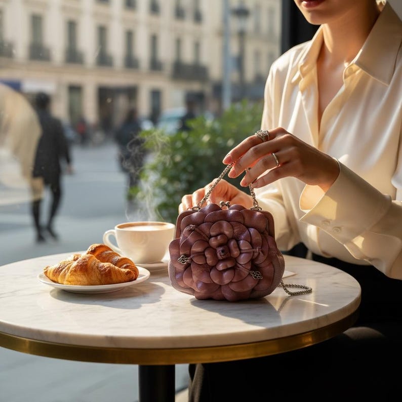 Puede incluir: Un bolso de mano de cuero malva con dise&ntilde;o floral y cadena plateada. El bolso est&aacute; sobre una mesa de m&aacute;rmol, junto a una taza de caf&eacute; y cruasanes. La escena tiene lugar en un caf&eacute;.