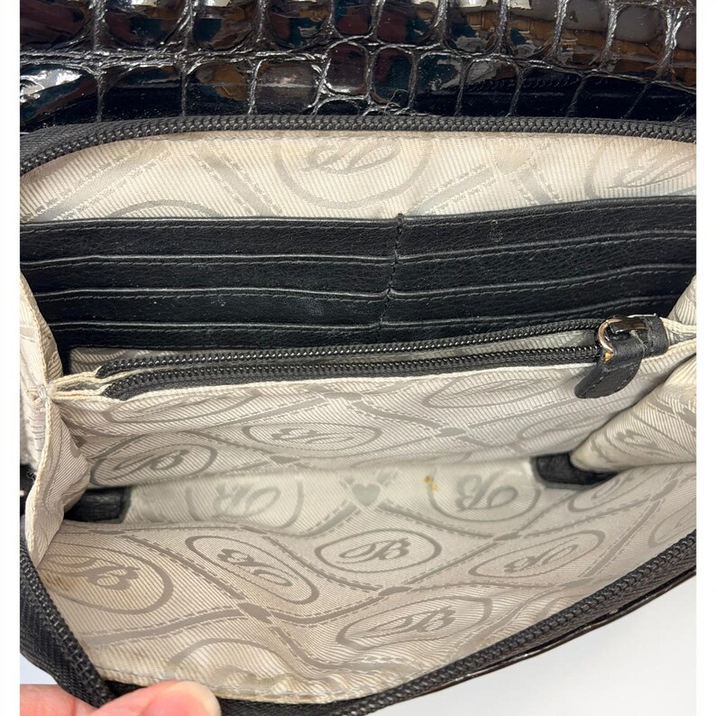 May include: A black wallet with a glossy crocodile-embossed exterior flap and a light-colored interior lining featuring a repeating logo pattern. The interior includes multiple card slots and a central zippered compartment with a black metal pull.