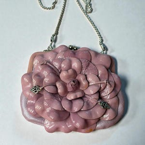 Puede incluir: Bolso de mano de cuero rosa claro con forma de flor de camelia. El bolso tiene una correa de cadena plateada y un cierre de boquilla. Acentos decorativos plateados visibles en los p&eacute;talos. Un accesorio &uacute;nico.