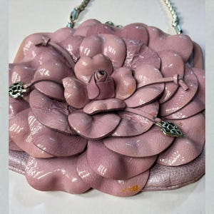 Puede incluir: Bolso de mano de cuero rosa claro con forma de flor de camelia. El bolso presenta una correa de cadena plateada y herrajes decorativos plateados. Los p&eacute;talos son texturizados y en capas, con un peque&ntilde;o capullo de rosa en el centro. Un accesorio &uacute;nico y elegante.