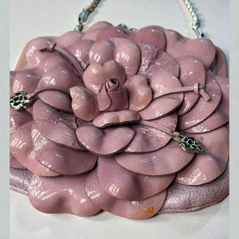 Puede incluir: Bolso de mano de cuero rosa claro con forma de flor de camelia. El bolso presenta una correa de cadena plateada y herrajes decorativos plateados. Los p&eacute;talos son texturizados y en capas, con un peque&ntilde;o capullo de rosa en el centro. Un accesorio &uacute;nico y elegante.