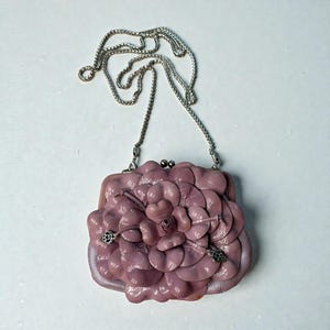 Puede incluir: Un bolso de mano de color malva con un dise&ntilde;o floral. El bolso tiene una correa de cadena plateada y un cierre de broche. El dise&ntilde;o floral es tridimensional, con m&uacute;ltiples capas de p&eacute;talos. El bolso est&aacute; hecho de un material suave, posiblemente cuero.