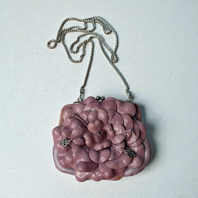 Puede incluir: Un bolso de mano de color malva con un dise&ntilde;o floral. El bolso tiene una correa de cadena plateada y un cierre de broche. El dise&ntilde;o floral es tridimensional, con m&uacute;ltiples capas de p&eacute;talos. El bolso est&aacute; hecho de un material suave, posiblemente cuero.