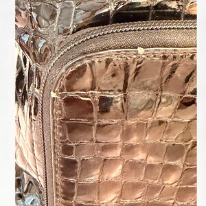 May include: A dark brown bag with a glossy, patent finish and a textured crocodile skin pattern. The bag features a prominent zipper closure along the top edge, with visible stitching detail along the seams of the structured, rectangular compartment.