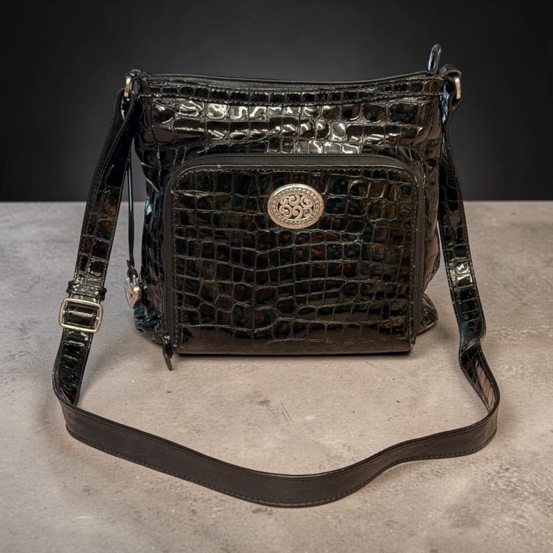 May include: A black crossbody bag with a glossy, embossed crocodile-skin texture. It features a front zippered pocket adorned with a central oval silver-toned metal emblem with intricate scrollwork and an adjustable shoulder strap with metal hardware.
