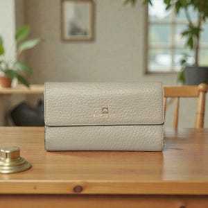 May include: A light gray leather wallet with a textured surface. The wallet has a flap closure and a gold logo. The wallet is sitting on a wooden table.