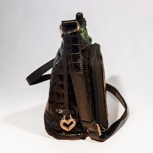 May include: A dark brown, glossy, crocodile-embossed shoulder bag with a structured silhouette. The bag features a side zippered pocket, a thin adjustable strap, and a metallic heart-shaped charm attached to the base of the strap hardware.