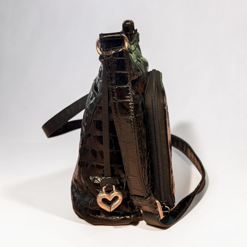 May include: A dark brown, glossy, crocodile-embossed shoulder bag with a structured silhouette. The bag features a side zippered pocket, a thin adjustable strap, and a metallic heart-shaped charm attached to the base of the strap hardware.