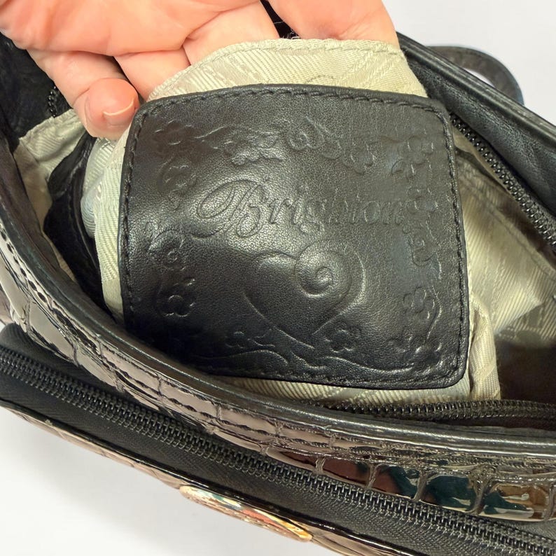 May include: A black handbag featuring a rectangular leather patch embossed with the word Brighton, a heart icon, and floral motifs. The bag has a glossy, textured exterior with a crocodile-like pattern and a zippered closure with light fabric lining.