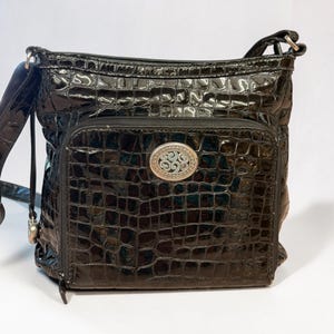 May include: A black crossbody bag with a glossy, embossed crocodile skin texture. The bag features a front zippered pocket adorned with an oval silver-toned metal emblem containing an intricate, swirling filigree design in the center.