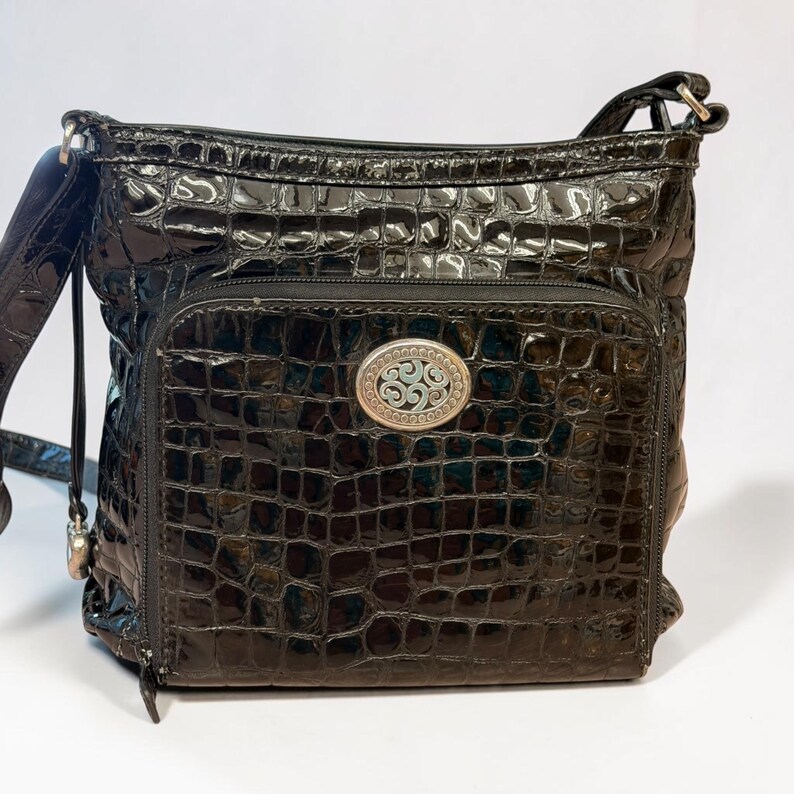 May include: A black crossbody bag with a glossy, embossed crocodile skin texture. The bag features a front zippered pocket adorned with an oval silver-toned metal emblem containing an intricate, swirling filigree design in the center.