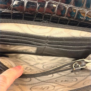 May include: A black wallet with a glossy crocodile-embossed texture on the exterior flap. The interior features a light beige fabric lining with a repeating logo pattern, multiple horizontal card slots, and a zippered compartment for storage.