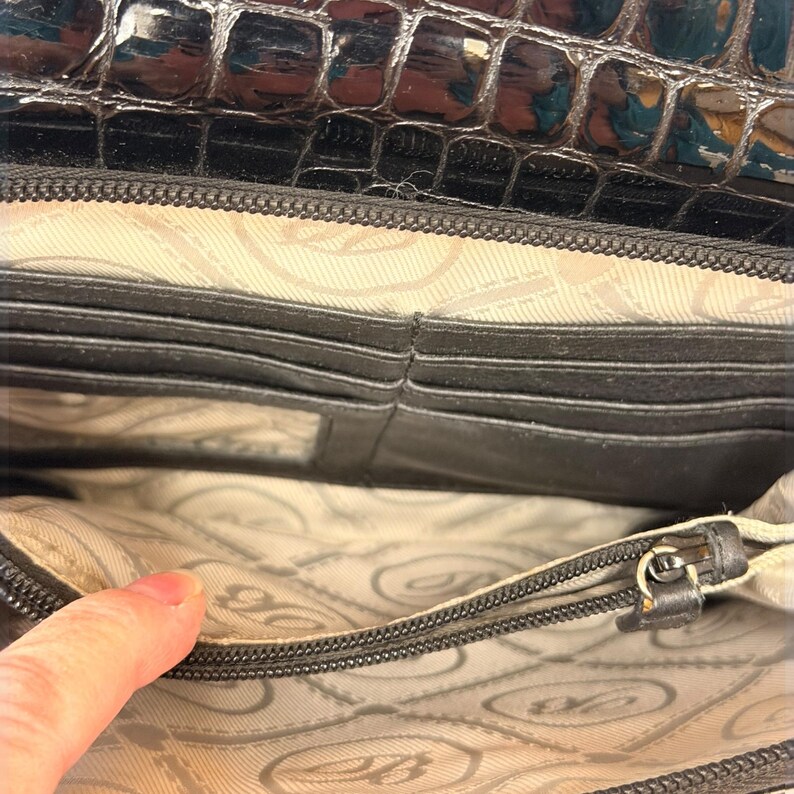 May include: A black wallet with a glossy crocodile-embossed texture on the exterior flap. The interior features a light beige fabric lining with a repeating logo pattern, multiple horizontal card slots, and a zippered compartment for storage.