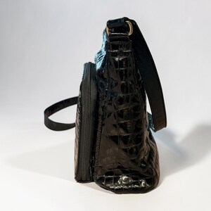 May include: A black shoulder bag with a glossy, embossed crocodile skin texture. The bag features a structured rectangular shape, a side zipper compartment, and a matching black shoulder strap attached with metallic hardware on the top side.
