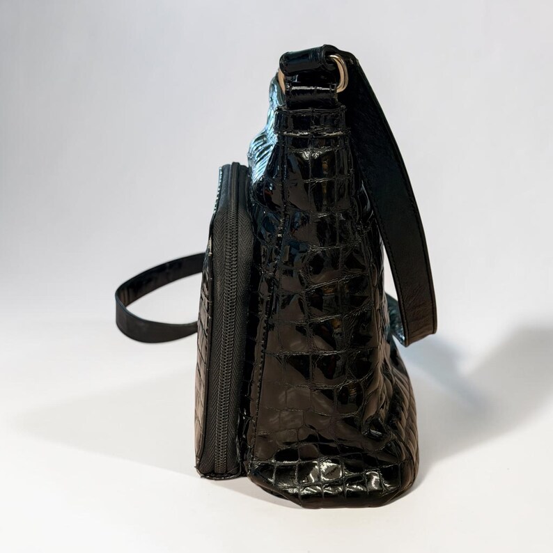 May include: A black shoulder bag with a glossy, embossed crocodile skin texture. The bag features a structured rectangular shape, a side zipper compartment, and a matching black shoulder strap attached with metallic hardware on the top side.