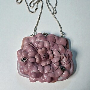Puede incluir: Un bolso de mano de cuero lavanda con un dise&ntilde;o floral. El bolso presenta una correa de cadena plateada y un cierre de broche. El dise&ntilde;o floral es tridimensional, con m&uacute;ltiples capas de p&eacute;talos. Fondo blanco.