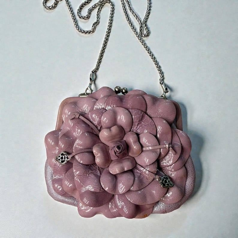 Puede incluir: Un bolso de mano de cuero lavanda con un dise&ntilde;o floral. El bolso presenta una correa de cadena plateada y un cierre de broche. El dise&ntilde;o floral es tridimensional, con m&uacute;ltiples capas de p&eacute;talos. Fondo blanco.