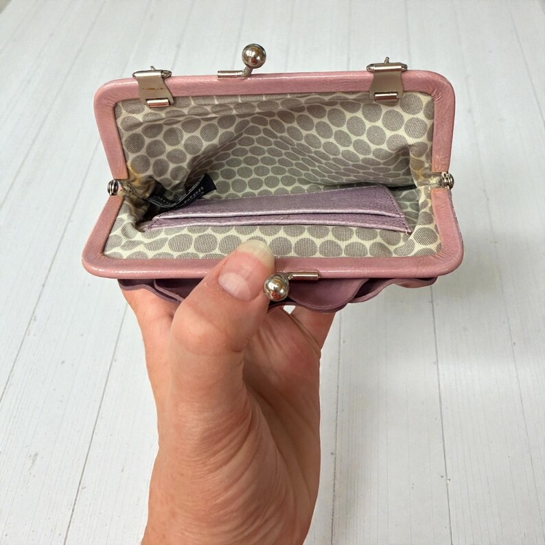 Puede incluir: Monedero rosa claro con cierre de beso abierto, con un dise&ntilde;o floral. El interior presenta un patr&oacute;n de lunares grises y blancos y un peque&ntilde;o bolsillo. El bolso est&aacute; abierto, revelando un peque&ntilde;o tarjetero.
