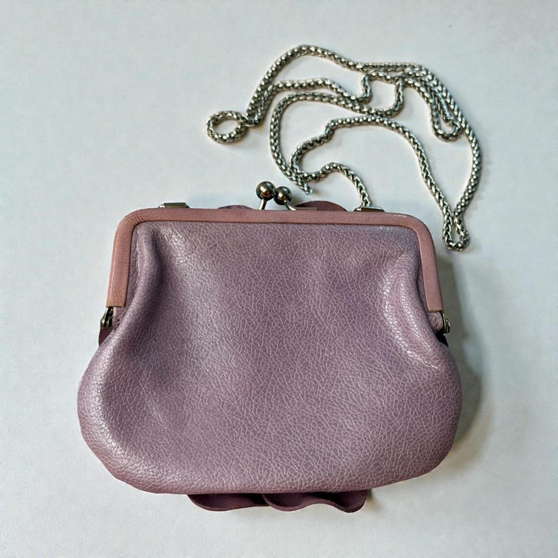 Puede incluir: Un monedero de cuero lavanda con una cadena plateada. El monedero tiene un cierre de boquilla y un detalle floral decorativo en la parte inferior. La cadena mide aproximadamente 50 cm de largo.