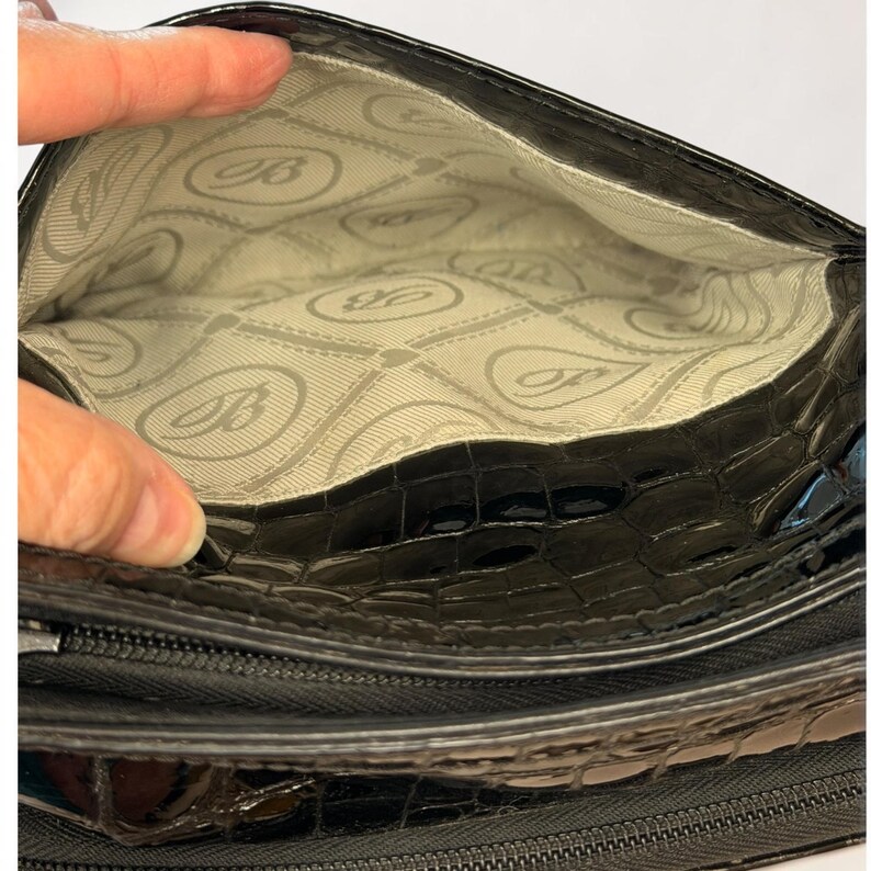 May include: A black handbag with a glossy, textured crocodile-embossed exterior. The interior is lined with a light-colored fabric featuring a repeating pattern of the letter B inside an oval shape, separated by thin, dashed horizontal lines.