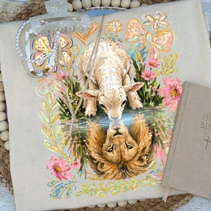 Può includere: Una t-shirt beige con la grafica di un agnello che guarda nell'acqua e vede il riflesso di un leone. Il design include fiori rosa, un fiocco dorato e foglie verdi. Sono presenti un fermaglio per capelli bianco, una collana di perle e una Bibbia.