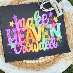 以下が含まれることがあります： 「make HEAVEN crowded」というカラフルな文字がレインボーグラデーションのフォントで描かれたダークグレーのTシャツ。ペンキが飛び散ったようなエフェクトと小さな星のグラフィックが特徴。白いビーズのブレスレットと半透明の白いヘアクリップが上に置かれている。