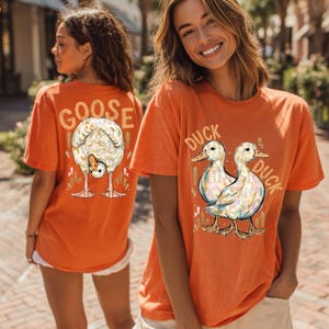 May include: Orange t-shirts with animal graphics. One shirt features a goose bending over with the word "GOOSE" above it. The other shirt has two ducks and the words "DUCK DUCK". The shirts are short-sleeved and have a casual style.