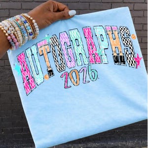 Op de afbeelding: Lichtblauw T-shirt met het woord "AUTOGRAPHS" in een kleurrijk, patroonlettertype en het nummer "2026" eronder. Het shirt wordt omhooggehouden door een hand met armbanden.