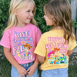 Peut inclure: Un t-shirt rose avec un graphique de Woody et le texte "just remember what your old pal said" et un t-shirt jaune avec un graphique de Jessie, Woody et Buzz l'Éclair avec le texte "YOU'VE GOT A FRIEND IN ME".
