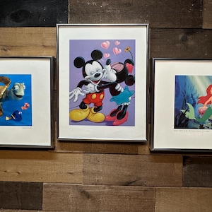 May include: Three framed Disney art prints. One features a turtle and fish in blue water, another shows Mickey Mouse and Minnie Mouse embracing, and the third depicts Ariel the mermaid with sea creatures.