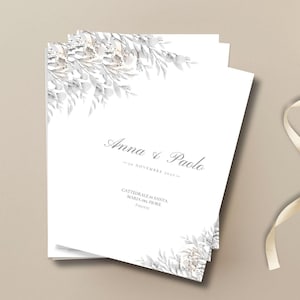 May include: White wedding invitation cards with floral and leaf accents. The cards feature the names "Anna & Paolo" and event details. A cream-colored ribbon and floral elements are in the background.