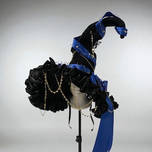 May include: A black witch hat with a wide brim and a tall, pointed crown. The hat is adorned with blue satin ribbons, gold star-shaped charms, and black ruffled fabric. A long blue ribbon drapes down the side.