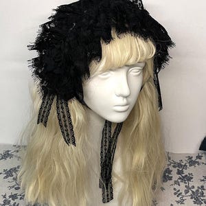 May include: A black lace bonnet featuring intricate floral patterns, ruffled edges, and delicate fabric ribbons that tie under the chin. The headpiece is displayed on a mannequin head with long blonde hair, showcasing the detailed lace texture.