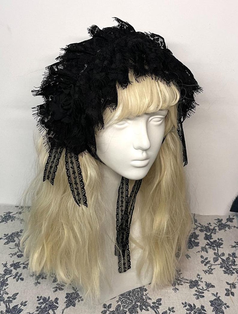 May include: A black lace bonnet featuring intricate floral patterns, ruffled edges, and delicate fabric ribbons that tie under the chin. The headpiece is displayed on a mannequin head with long blonde hair, showcasing the detailed lace texture.