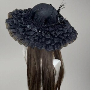 May include: A black hat with a wide, ruffled brim and a rounded crown. The hat is adorned with feathers and a decorative flower. The brim is made of a sheer, layered fabric, creating a voluminous effect. The hat is displayed on a mannequin head.