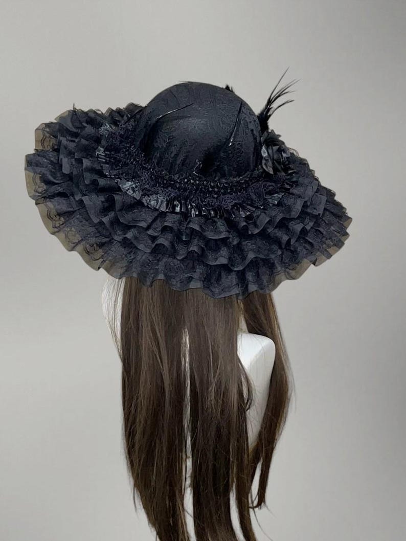 May include: A black hat with a wide, ruffled brim and a rounded crown. The hat is adorned with feathers and a decorative flower. The brim is made of a sheer, layered fabric, creating a voluminous effect. The hat is displayed on a mannequin head.