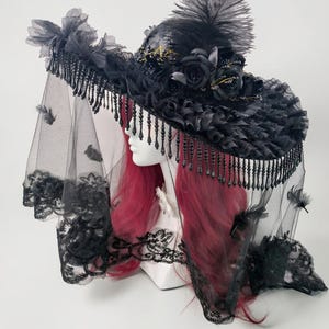 Puede incluir: Un sombrero negro de ala ancha adornado con rosas de tela negra, un penacho de plumas altas y volantes. Un velo negro largo y transparente con bordes de encaje y peque&ntilde;as plumas dispersas cuelga del ala, decorada con cuentas negras colgantes.