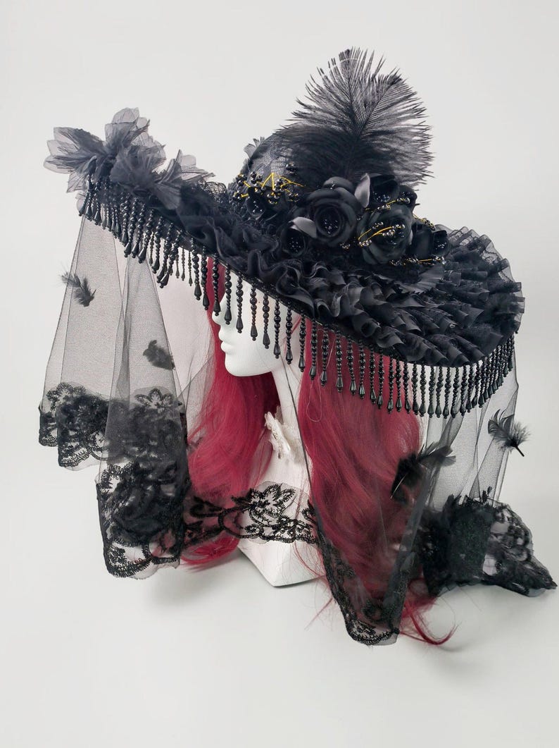 Puede incluir: Un sombrero negro de ala ancha adornado con rosas de tela negra, un penacho de plumas altas y volantes. Un velo negro largo y transparente con bordes de encaje y peque&ntilde;as plumas dispersas cuelga del ala, decorada con cuentas negras colgantes.
