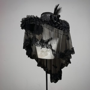 Puede incluir: Un sombrero negro de ala ancha con encaje, un grupo de rosas de tela negra y plumas erguidas. El sombrero est&aacute; adornado con cuentas negras colgantes a lo largo del ala y un velo negro largo y transparente con bordes de encaje intrincados.