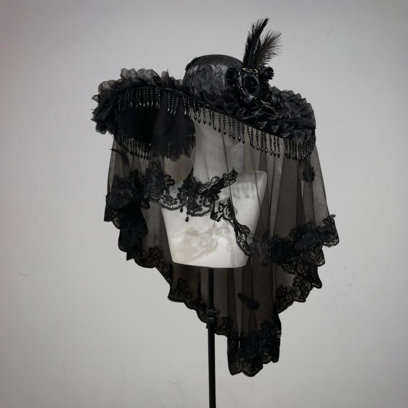 Puede incluir: Un sombrero negro de ala ancha con encaje, un grupo de rosas de tela negra y plumas erguidas. El sombrero est&aacute; adornado con cuentas negras colgantes a lo largo del ala y un velo negro largo y transparente con bordes de encaje intrincados.